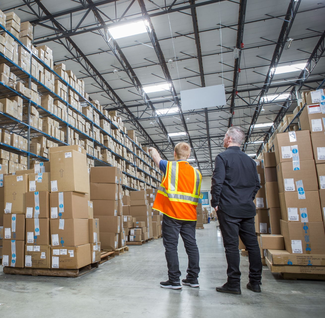 Man in hi-vis vest pointing to boxes while man in black shirt looks in warehouse
